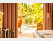 Blick aus einem Zimmer mit gestreifter Tapete und Holzmöbeln auf eine Terrasse mit einer Hängematte und einem Pool, umgeben von tropischen Pflanzen. Im Hintergrund sind Menschen im Wasser zu sehen.