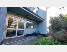 Eingangsbereich eines einfachen Gebäudes mit blauer Fassade. Vor der verglasten Tür mit der Hausnummer 10 steht ein Fahrrad auf einem gepflasterten Boden, umgeben von Gebüsch und einer Hecke; im Hintergrund sind weitere Gebäude zu sehen.