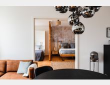 Wohnbereich mit braunem Sofa, schwarzem Esstisch und einer modernen, spiegelnden Hängelampe im Vordergrund. Durch eine Türöffnung ist ein Schlafzimmer mit Holzboden, zwei Betten, einer Tapete mit floralem Muster und warmen Holzmöbeln zu sehen.