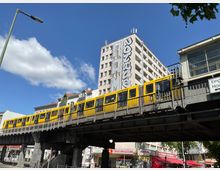 Gelbe U-Bahn auf einer erhöhten Bahntrasse in einer urbanen Umgebung, im Hintergrund ein mehrstöckiges Gebäude mit großem Graffiti. Unten befinden sich Geschäfte und Schilder, die auf Supermärkte hinweisen.