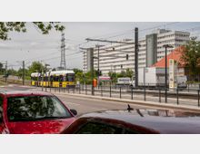 Straßenszene mit einer gelb-weißen Straßenbahn an einer Haltestelle, umgeben von Verkehrsschildern, parkenden Autos und Gehwegen. Im Hintergrund sind ein großes Wohngebäude, ein Strommast und grüne Bäume zu sehen.