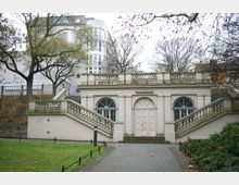 Historisches Gebäude mit symmetrischer Architektur, bestehend aus einem zentralen weißen Eingang mit Doppeltür, flankiert von Bögen mit vergitterten Fenstern und Treppen auf beiden Seiten. Im Vordergrund befindet sich ein gepflasterter Weg, Rasenflächen und Bäume, während modernere Gebäude im Hintergrund sichtbar sind.