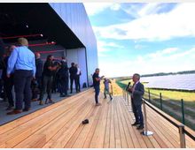 Eine moderne Außenveranstaltung auf einer hölzernen Terrasse vor einem großen Gebäude mit einer geöffneten Seite. Im Hintergrund sind weitläufige Solarpanels auf einer Wiese unter blauem Himmel zu sehen.