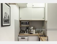 Kleine, moderne Kochnische mit weißen Oberschränken, einer Arbeitsplatte aus Holz und Geräten wie einer Mikrowelle, einer Kaffeemaschine und einer Spülmaschine. An der Wand hängt ein gerahmtes Schwarz-Weiß-Foto, daneben befinden sich eine Spüle und weitere Küchenutensilien.