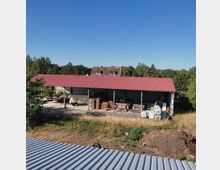 Eine Lagerhalle mit rotem Satteldach und offenen Seiten steht auf einem Gelände mit gestapelten Holzpaletten, Baumaterialien und einem geparkten Transporter. Im Hintergrund sind ein Wohnhaus mit Dachgauben und ein bewaldeter Bereich zu sehen, umgeben von Wiesen und Büschen.