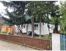 Weißes Einfamilienhaus mit flachem Dach, umgeben von hohen Bäumen in einem grünen Garten. Der Garten ist von einem Holz- und Metallzaun eingefasst, im Vordergrund Pflasterweg und straßenseitige Ansicht.