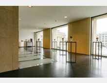 Ein moderner Innenraum mit großen Fensterfronten, die Ausblick auf das Brandenburger Tor bieten. Der Raum ist mit mehreren hohen Stehtischen ausgestattet, die jeweils mit kleinen Blumenvasen dekoriert sind, und verfügt über einen dunklen Boden sowie beige Wände. Im Hintergrund ist eine Kaffeemaschine auf einer Theke erkennbar.