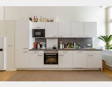 Moderne Einbauküche mit weißen Schränken und grauer Arbeitsplatte. In der Küche befinden sich ein Backofen, eine Mikrowelle, eine Kaffeemaschine und verschiedene Gewürze; ein großes Fenster und eine Zimmerpflanze ergänzen den Raum.