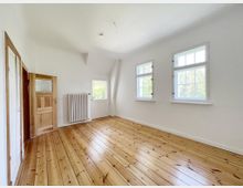 Heller, leerer Raum mit Holzdielenboden, weiß gestrichenen Wänden und drei Fenstern, die viel Tageslicht hereinlassen. Links im Raum befinden sich eine Holztür und ein weißer Heizkörper.