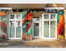 Eingangsbereich eines Wohn- oder Geschäftsgebäudes mit weiß gerahmter Glastür und großen Fenstern, umgeben von einer bunt bemalten Graffiti-Fassade. Vor dem Eingang liegt ein gepflasterter Gehweg, in den Scheiben spiegeln sich Bäume und die Straße.