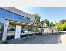 Ein einstöckiges Gebäude mit großen Glasfronten und geschwungener Beschriftung "L'Aiglon" auf dem Dachvorsprung. Die Umgebung ist von Bäumen gesäumt, und auf dem Vorplatz stehen geparkte Autos.