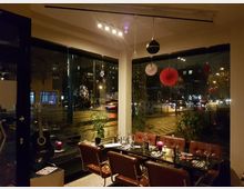 Ein gemütliches Restaurant mit einer langen Tafel, gedeckt mit Gläsern und Besteck. Große Fensterfronten zeigen eine nächtliche Straßenansicht mit vorbeifahrenden Autos und Dekorationen wie rote Papierornamente und Lichtreflexionen auf dem Glas.