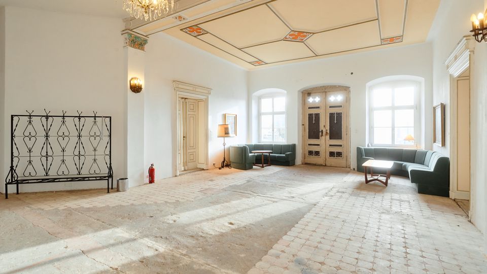 Großzügiger, heller Innenraum mit weiß verputzten Wänden, hohen Fenstern und einer doppelflügeligen Eingangstür. Auffällig sind die dekorierte Decke mit Kronleuchter, der abgenutzte Fliesenboden sowie Sitzgruppen mit grünen Sofas und kleinen Tischen entlang der Wände.