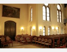 Innenraum einer historischen Kirche mit hellbeigen Wänden, hohen Bogenfenstern mit Buntglas und Wandlampen, die warmes Licht spenden. Holzbänke und gepolsterte Stühle stehen entlang der Wände, über einem dunklen Holztor hängt ein großes Gemälde.