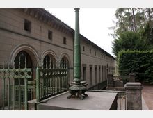 Historisches Gebäude mit einer Reihe von Bogenfenstern und einer Fassade aus grauem Stein, umgeben von einem grünen, dekorativen Metallzaun. Im Vordergrund steht eine kunstvoll verzierte Metalllaterne auf einem steinernen Sockel, rechts ist dichter Bewuchs zu sehen.