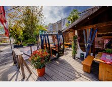 Holzterrasse eines rustikalen Lokals mit Holzbänken und Tischen, bepflanzten Kübeln und einer dekorativen Gondelkabine als Sitzbereich. Daneben eine hölzerne Hütte mit an der Wand befestigten Skiern; im Hintergrund Bäume sowie Gebäude mit Graffiti und Baustellenkran.