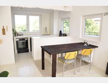 Heller Wohnraum mit offener Küche und Essbereich. Die Küche verfügt über weiße Wände, große Fenster mit Blick ins Grüne, eine Arbeitsplatte mit Spüle und einen modernen Backofen; im Vordergrund steht ein dunkler Holztisch mit gelben Stühlen auf einem hellen Teppich.