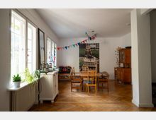 Heller Wohn- und Essbereich mit großen Fenstern entlang der linken Wand, Holzboden im Fischgrätmuster und einem Tisch mit Stühlen in der Raummitte. An der Rückwand hängt ein großes Wandbild, darüber eine bunte Wimpelgirlande; rechts steht ein Holzsideboard mit Pflanzen.