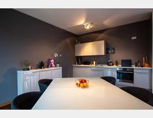 Moderne Küche mit grauen Wänden und einer Kombination aus weißen Unterschränken und Hängeschränken. Im Vordergrund steht ein Esstisch mit glänzender weißer Oberfläche und einem Obstkorb, umgeben von dunklen Stühlen; die Arbeitsplatte ist mit Küchengeräten und Dekorationsgegenständen bestückt.