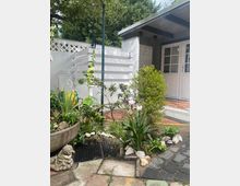 Ein kleiner Garten mit dichtem Grün, dekorativen Pflanzen und Blüten, umgeben von Pflastersteinen und Ziegeln. Im Hintergrund ein weiß gestrichenes Gebäude mit Holztüren und einem Flachdach, daneben eine Außendusche mit Sichtschutzwand.