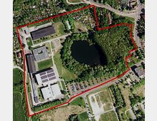 Luftbild einer großen, umzäunten Fläche mit Gebäuden im linken Bereich, einem zentralen Teich, umgeben von dichtem Grün und Parkplätzen entlang der Straße. Im oberen Bereich sind Kleingärten und benachbarte Straßen zu erkennen.