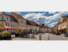 Eine gepflasterte Altstadtstraße mit historischen Gebäuden in Pastellfarben und roten Ziegeldächern. Links stehen Cafés mit roten Sonnenschirmen, Fahrräder sind abgestellt, und Blumenampeln schmücken Straßenlaternen.