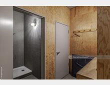 Holzvertäfelter Umkleideraum mit Hakenleiste und Bank; links ist eine geöffnete Tür, die den Zugang zu einer kleinen, mit grauen Fliesen ausgestatteten Duschkabine zeigt.