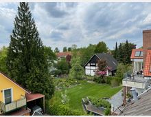 Blick auf einen grünen, gepflegten Garten mit Rasenfläche, umgeben von mehreren Einfamilienhäusern, darunter ein gelbes Haus mit Balkon, ein Fachwerkhaus mit weißen Wänden und ein rotes Backsteinhaus mit Terrasse. Im Hintergrund sind hohe Bäume und bewölkter Himmel sichtbar.