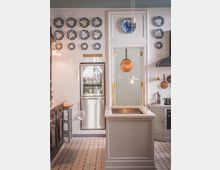 Küche mit heller Wandverkleidung aus weißen Fliesen, dekoriert mit blau-weißen Ziertellern. Zentral steht eine graue Kücheninsel mit Marmorarbeitsplatte, umgeben von klassischen Küchenelementen wie einem Holzschrank, einem Edelstahlkühlschrank und einem Herd.