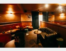 Gemütlicher Lounge- oder Bar-Innenraum mit gepolsterten Sitzbänken in Rot und einem schwarzen Chesterfield-Sofa, runden Marmortischen und dunklem Fliesenboden. Warme Wand- und Deckenbeleuchtung, Spiegelwand sowie Vorhänge und Wandventilatoren prägen den Raum.