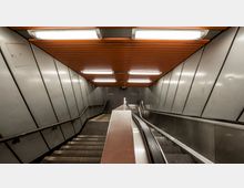 Treppenabgang und Rolltreppe in einer U-Bahn-Station mit grauen Metallwänden und einer Decke aus orangefarbenen Holzpaneelen, beleuchtet durch mehrere rechteckige Leuchtstofflampen.