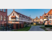 Backsteingebäude in einer historischen Wohnanlage mit spitz zulaufenden Giebeln und roten Ziegeldächern, umgeben von einem gepflasterten Weg, grünen Rasenflächen und einigen Bäumen; im Vordergrund ein Fahrradständer, im Hintergrund ein weiteres Gebäude mit neogotischen Elementen.