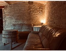 Gemütlicher Raum mit freiliegenden Backsteinwänden, zwei braunen Ledersofas, einem rustikalen Holztisch und einer kleinen Lampe auf einem runden, weißen Tisch. Die Decke ist gewölbt und der Raum wird durch warmes Licht beleuchtet.