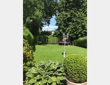 Ein gepflegter Garten mit einer Rasenfläche, umgeben von hohen Hecken und Bäumen; im Hintergrund eine Schaukel und ein Trampolin. Im Vordergrund befinden sich dekorative Sträucher und eine Hostapflanze, mit einer Laterne auf der Rasenfläche.