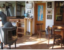 Wohnzimmer mit rustikalem Stil: Ein freistehender schwarzer Kaminofen steht im Vordergrund, daneben Holzmöbel, darunter ein Schrank, Stühle und eine kleine Leiter. An der Wand hängen Bilder, ein Wandposter einer Karte und ein Globus steht auf einem Holzregal.