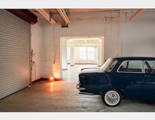 Innenraum einer industriellen Lager- oder Werkstatthalle mit Betonboden, weiß gestrichenen Wänden, sichtbaren Leitungen und einer großen Rolltor-Tür links. Durch eine breite Öffnung führt der Blick in einen hellen Nebenraum mit großen Fenstern; rechts ist ein teilweise sichtbares dunkelblaues Auto geparkt.