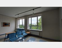 Wartebereich mit blauen Sitzbänken und kleinen Tischen vor großen Fenstern, die eine Aussicht auf Bäume und Gebäude im Hintergrund bieten. Die beige gestrichenen Wände sind mit einem Bild dekoriert, und eine Deckenleuchte ist mittig angebracht.