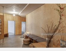 Minimalistisch gestalteter Innenraum mit Wänden aus hellem Sperrholz, einer Bank mit grauen Kissen, runden geflochtenen Sitzhockern und einem Tisch mit einer Vase, die Zweige mit Blüten enthält. Im Hintergrund führen offene Türrahmen zu angrenzenden Räumen in schlichtem, modernem Stil.