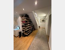 Flur in einem Wohnhaus mit hellem Holzfußboden und einer langen grauen Läufermatte. Links befindet sich ein großes Regal mit mehreren Ablagen voller Schuhe, daneben ein kleiner weißer Schrank. Geradeaus führt eine weiße Treppe nach oben, an der Wand hängt ein gerahmtes Bild. Deckenspots sorgen für Beleuchtung.