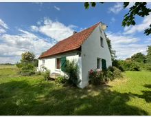 Weißes Landhaus mit rotem Ziegeldach und grünen Fensterläden, umgeben von einer Wiese und Bäumen. Im Vordergrund sind Büsche, Blumen und eine Holzbank, im Hintergrund eine weitläufige Landschaft unter leicht bewölktem Himmel zu sehen.