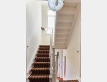 Treppe in einem Wohnhaus mit gemustertem Teppichbelag, weißen Wänden und einem Holzgeländer. Eine kleine Fensterbank mit Dekoration und ein Fenster im Hintergrund lassen Tageslicht in den Raum.