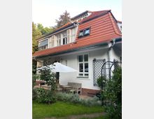 Ein Einfamilienhaus mit weißer Fassade, roten Dachziegeln und mehreren Fenstern, umgeben von einem Garten mit Pflanzen und einer kleinen Terrasse. Die Terrasse ist mit einem weißen Sonnenschirm, Holzmöbeln und Stühlen ausgestattet.