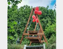 Holzschaukel mit Kettenaufhängung unter einem A-förmigen Holzgestell, geschmückt mit roten Luftballons, vor üppigem grünen Laub im Freien.