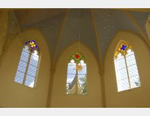 Innenraum einer Kirche mit drei gotischen Spitzbogenfenstern, verziert mit Buntglas in Blau, Grün, Rot und Gelb. Die Decke ist himmelblau mit goldenen Sternen verziert.