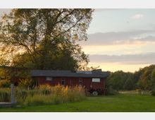 Ein rotbrauner, rustikaler Bauwagen steht auf einer Wiese, umgeben von hohem Gras und Bäumen, im Hintergrund ein ländliches Waldgebiet. Der Himmel zeigt eine Abendstimmung mit weichen, orangefarbenen und blauen Wolkentönen.