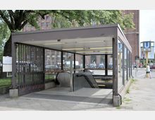 Eingang zu einer U-Bahn-Station mit einer Überdachung aus Glas und Metall sowie seitlichen Gitterwänden. Im Vordergrund eine Rolltreppe und Treppen, im Hintergrund eine Straße mit Autos, Fußgängern und einem Backsteingebäude.