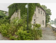 Ein älteres, verputztes Gebäude, teils von dichtem Efeu überwuchert, mit mehreren einfachen Fenstern. Vor dem Gebäude stehen Büsche und ein gepflasterter Weg führt entlang eines angrenzenden Flusses.