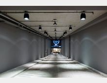 Langer, schmaler Innenraum mit grauen Wänden und einer Betondecke, die mit schlichten schwarzen Deckenleuchten ausgestattet ist. Am Ende des Gangs befindet sich ein heller Raum mit Möbeln und eine beleuchtete Wand mit einer dekorativen Projektion.