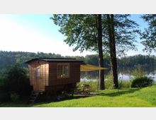 Holzwagen auf Rädern mit kleinem Vorbau und gelber Markise, umgeben von grüner Wiese und Bäumen am Ufer eines ruhigen Sees mit bewaldetem Hintergrund.