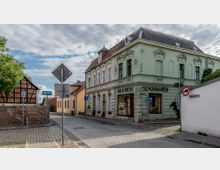 Straßenszene in einem historischen Viertel mit gepflasterten Wegen; rechts ein pastellgrünes Gebäude mit der Aufschrift „Wilh. Riese Schuhwaren“ und dekorativen Fassadenelementen, links ein Fachwerkhaus aus roten Ziegeln. Straßenschilder und ein Baum prägen den Vordergrund.